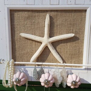 May include: A white wooden frame with a burlap background and a white starfish. The frame has a white metal rod with pink ceramic knobs and necklaces and earrings hanging from it.