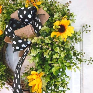 May include: A decorative wreath featuring bright yellow sunflowers and lush green foliage. A prominent bow made of black ribbon with white polka dots and tan burlap trim is attached to the side, adding a contrasting pattern to the floral arrangement.