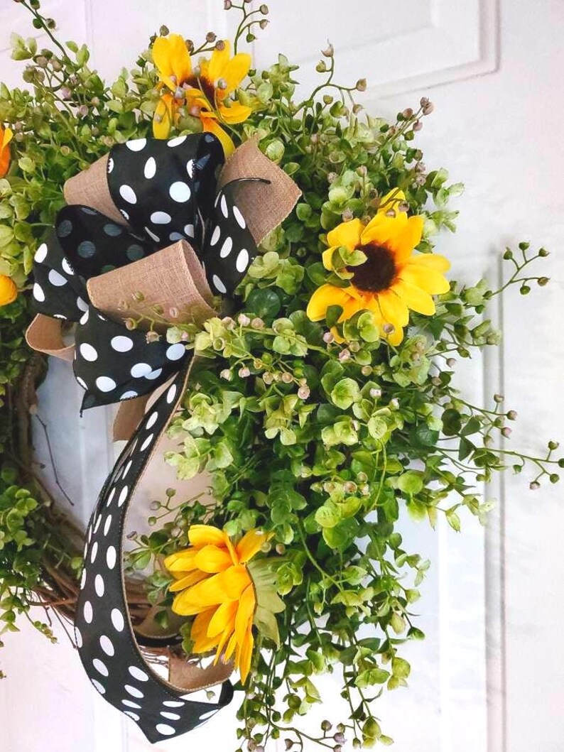 May include: A decorative wreath featuring bright yellow sunflowers and lush green foliage. A prominent bow made of black ribbon with white polka dots and tan burlap trim is attached to the side, adding a contrasting pattern to the floral arrangement.