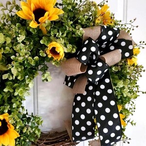 May include: A decorative wreath featuring bright yellow sunflowers and green foliage arranged on a circular twig base. A large, multi-layered bow made of black fabric with white polka dots and tan burlap ribbon is attached to the side of the wreath.