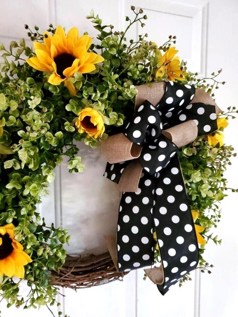 May include: A decorative wreath featuring bright yellow sunflowers and green foliage arranged on a circular twig base. A large, multi-layered bow made of black fabric with white polka dots and tan burlap ribbon is attached to the side of the wreath.