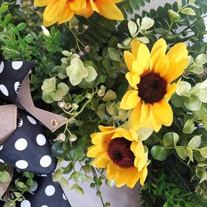 May include: A decorative wreath featuring three bright yellow sunflowers with dark brown centers, surrounded by lush green foliage and small sprigs. A black ribbon with white polka dots and a tan burlap accent is attached to the side of the arrangement.