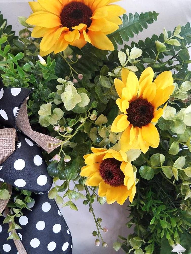 May include: A decorative wreath featuring three bright yellow sunflowers with dark brown centers, surrounded by lush green foliage and small sprigs. A black ribbon with white polka dots and a tan burlap accent is attached to the side of the arrangement.