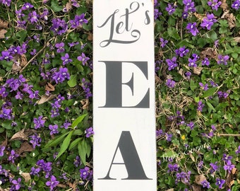 May include: A rectangular, white wooden sign with the words "Let's EAT" in black lettering. The sign is placed in a bed of purple flowers and green foliage, creating a rustic, outdoor aesthetic.