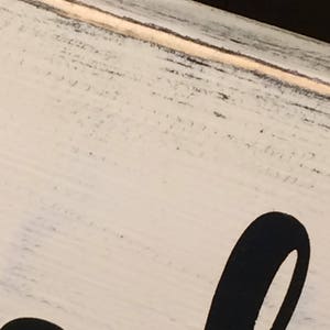 May include: Close-up of a distressed, off-white wooden sign with black lettering. The sign has a rustic, farmhouse style with visible wood grain and a slightly weathered appearance. The black lettering is large and stylized.