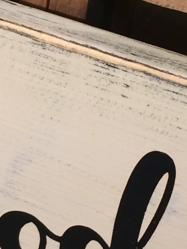 May include: Close-up of a distressed, off-white wooden sign with black lettering. The sign has a rustic, farmhouse style with visible wood grain and a slightly weathered appearance. The black lettering is large and stylized.