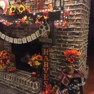 May include: A brick fireplace decorated for autumn with sunflowers, leaves, and a "Happy Fall" banner. A wooden sign reads "Harvest". A black lantern and fairy lights add to the festive atmosphere. A decorative owl is also present.