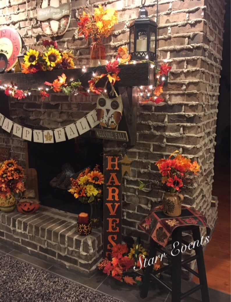 May include: A brick fireplace decorated for autumn with sunflowers, leaves, and a "Happy Fall" banner. A wooden sign reads "Harvest". A black lantern and fairy lights add to the festive atmosphere. A decorative owl is also present.