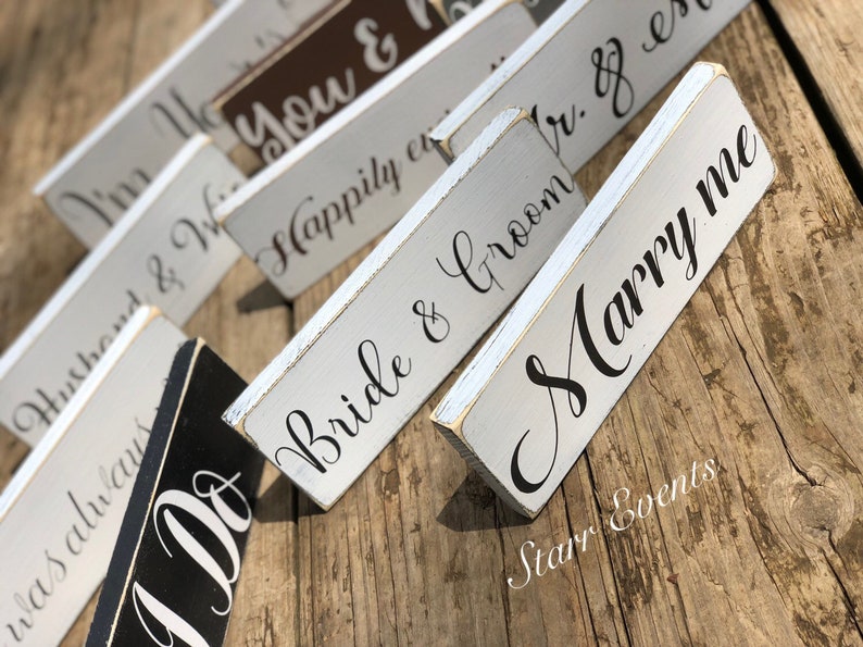May include: Wooden signs with white and black painted surfaces, featuring phrases like "Marry me", "Bride & Groom", and "I Do" in elegant script. The signs are arranged on a wooden surface, ideal for wedding decor.