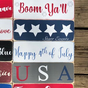 May include: A selection of rectangular wooden signs with patriotic themes. The signs display phrases such as "America," "Boom Ya'll," "Happy 4th of July," and "Freedom" in red, white, and blue. The signs are arranged on a wooden surface.
