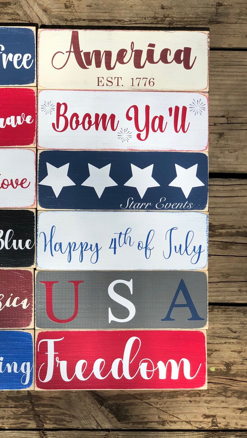 May include: A selection of rectangular wooden signs with patriotic themes. The signs display phrases such as "America," "Boom Ya'll," "Happy 4th of July," and "Freedom" in red, white, and blue. The signs are arranged on a wooden surface.