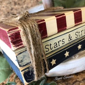 Puede incluir: Una pila de tres bloques de madera atados con cuerda, pintados con colores patri&oacute;ticos. El bloque superior tiene rayas rojas y blancas, el bloque del medio dice "Stars & Stripes", y el bloque inferior es azul con estrellas blancas. Una decoraci&oacute;n r&uacute;stica.