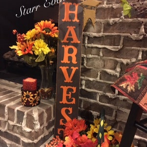 May include: A wooden sign with the word "HARVEST" in orange lettering. The sign is set against a brick wall and decorated with autumn flowers, a candle, and a star-shaped decoration.