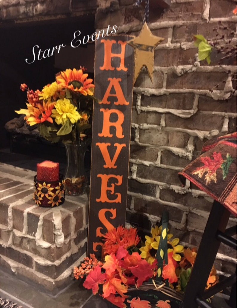 May include: A wooden sign with the word "HARVEST" in orange lettering. The sign is set against a brick wall and decorated with autumn flowers, a candle, and a star-shaped decoration.