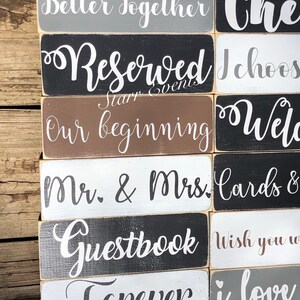 May include: A selection of rectangular wooden signs with white script lettering. Phrases include "Better Together," "Reserved," "Our Beginning," "Mr. & Mrs.," "Guestbook," and "Forever." The signs are painted in various colours, including grey, black, brown, and white.