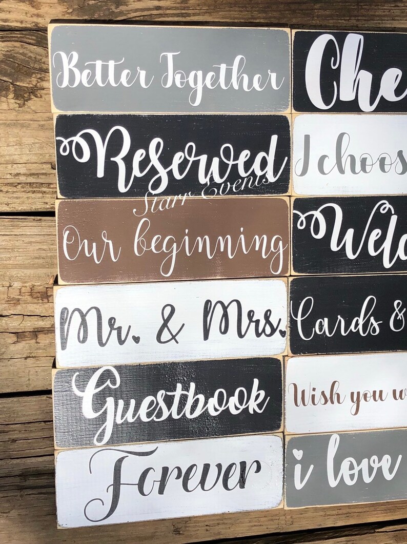 May include: A selection of rectangular wooden signs with white script lettering. Phrases include "Better Together," "Reserved," "Our Beginning," "Mr. & Mrs.," "Guestbook," and "Forever." The signs are painted in various colours, including grey, black, brown, and white.