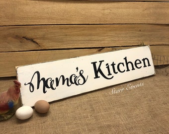 May include: A rustic, rectangular wooden sign with the words "Mama's Kitchen" in black script. The sign is painted white with distressed edges. A ceramic rooster and two eggs are in the foreground. The background is a wooden wall.