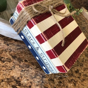 Puede incluir: Un conjunto de bloques de madera pintados con rayas rojas y blancas y azul con estrellas blancas. Los bloques est&aacute;n atados con hilo y presentan el texto "Stars & Stripes".