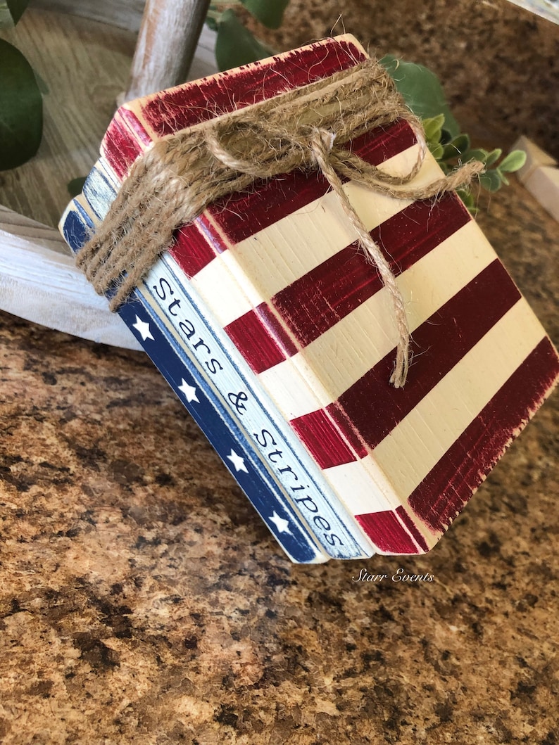 Puede incluir: Un conjunto de bloques de madera pintados con rayas rojas y blancas y azul con estrellas blancas. Los bloques est&aacute;n atados con hilo y presentan el texto "Stars & Stripes".