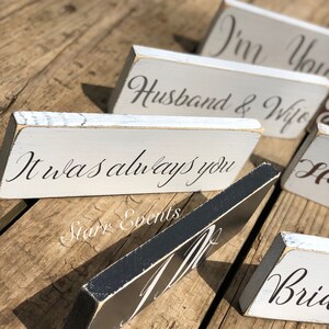 May include: Wooden signs with white and black painted surfaces and script lettering. The signs include phrases like "Husband & Wife" and "It was always you." The signs are arranged on a wooden surface.