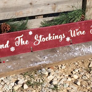 Pode incluir: Uma placa de madeira vermelha com o texto "And The Stockings Were Hung" em escrita branca, decorada com flocos de neve. A placa tem tr&ecirc;s ganchos para pendurar meias. Guirlanda de pinheiro e bagas vermelhas adornam a placa.