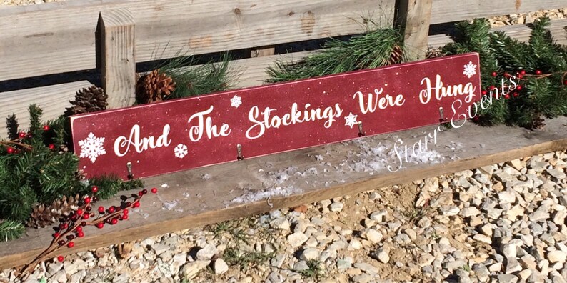 Pode incluir: Uma placa de madeira vermelha com o texto "And The Stockings Were Hung" em escrita branca, decorada com flocos de neve. A placa tem tr&ecirc;s ganchos para pendurar meias. Guirlanda de pinheiro e bagas vermelhas adornam a placa.
