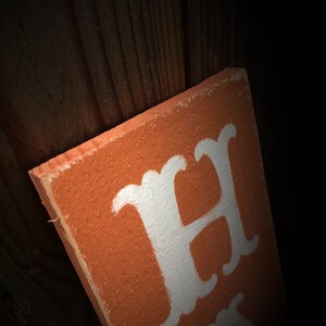 May include: Orange wooden sign with white lettering. The sign is angled, showing the edge and part of the text. The background is a dark wooden surface. The sign appears to be handmade.