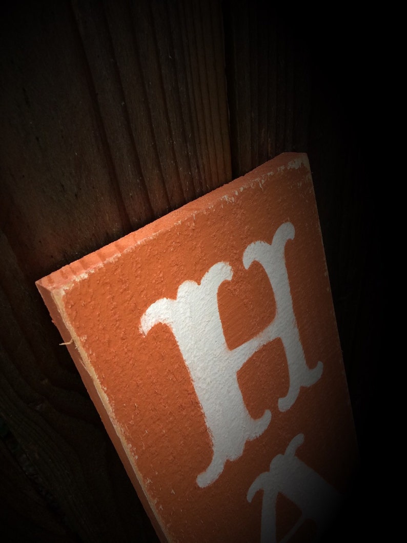 May include: Orange wooden sign with white lettering. The sign is angled, showing the edge and part of the text. The background is a dark wooden surface. The sign appears to be handmade.