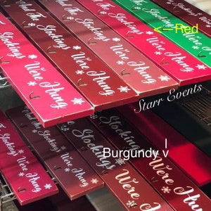 Pode incluir: Suportes de madeira para meias de Natal em v&aacute;rios tons de vermelho e verde. Cada suporte retangular tem o texto "And The Stockings Were Hung" e desenhos de flocos de neve. Inclui ganchos para pendurar meias.