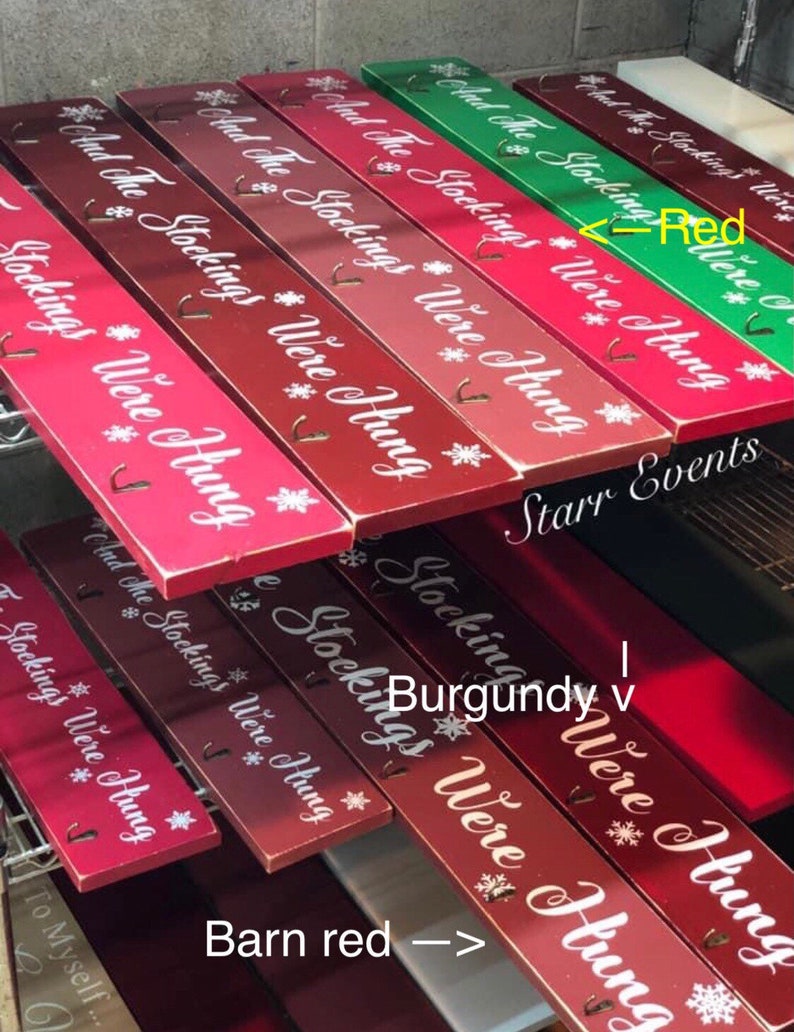 Pode incluir: Suportes de madeira para meias de Natal em v&aacute;rios tons de vermelho e verde. Cada suporte retangular tem o texto "And The Stockings Were Hung" e desenhos de flocos de neve. Inclui ganchos para pendurar meias.