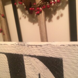 May include: Close-up of a distressed, white wooden sign with black lettering. The sign's edge is visible, showing a textured, weathered appearance. A wreath with red berries hangs in the background, adding a touch of color.
