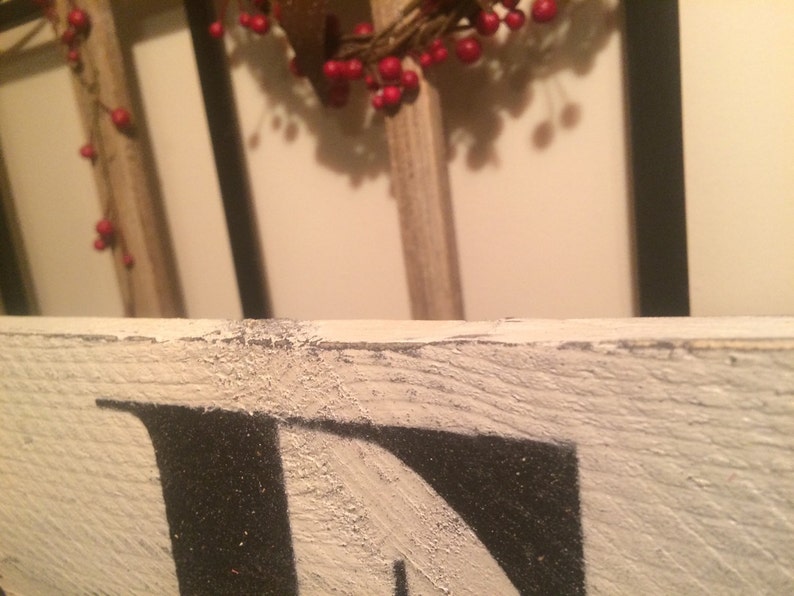 May include: Close-up of a distressed, white wooden sign with black lettering. The sign's edge is visible, showing a textured, weathered appearance. A wreath with red berries hangs in the background, adding a touch of color.