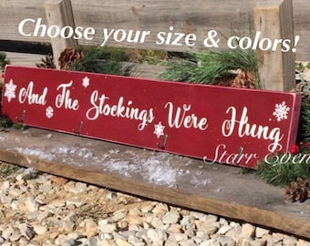 Op de afbeelding: Een rechthoekig, rood houten bord met de tekst "And The Stockings Were Hung" in wit script, geaccentueerd met sneeuwvlokken. Het bord bevat haken om kousen op te hangen. De tekst "Choose your size & colors!" staat bovenaan.