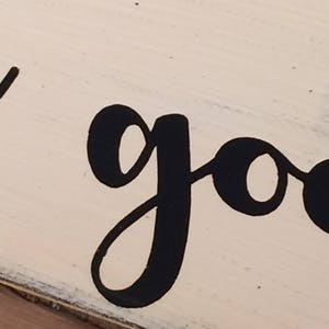 May include: Close-up of a wooden sign with the word "good" in large, black, cursive lettering. The sign has a distressed, off-white painted background and a dark border.