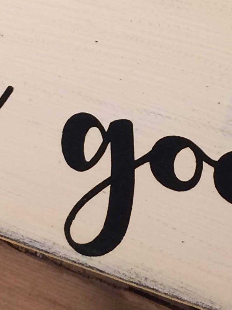 May include: Close-up of a wooden sign with the word "good" in large, black, cursive lettering. The sign has a distressed, off-white painted background and a dark border.