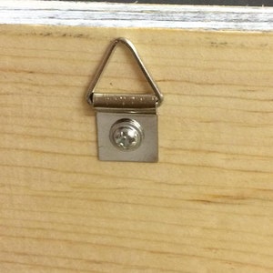 Pode incluir: Close-up de um painel de madeira com um cabide de metal para fotos fixado. O cabide tem formato de tri&acirc;ngulo prateado, fixado &agrave; madeira com uma pequena placa de metal e um &uacute;nico parafuso. O gr&atilde;o da madeira &eacute; vis&iacute;vel.