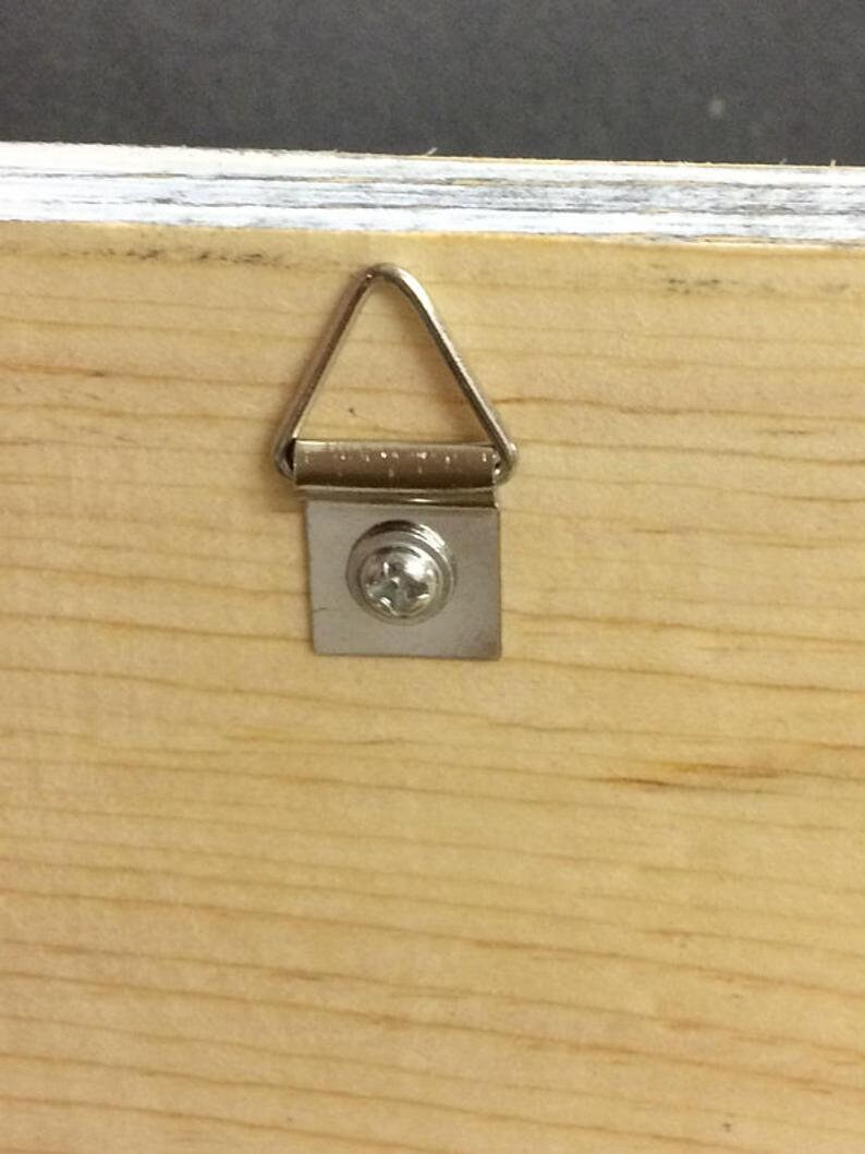 Pode incluir: Close-up de um painel de madeira com um cabide de metal para fotos fixado. O cabide tem formato de tri&acirc;ngulo prateado, fixado &agrave; madeira com uma pequena placa de metal e um &uacute;nico parafuso. O gr&atilde;o da madeira &eacute; vis&iacute;vel.