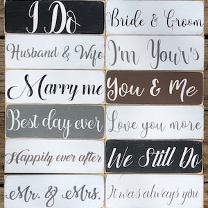 May include: A collection of rectangular wooden signs with white script. Phrases include "I Do", "Bride & Groom", "Husband & Wife", "Marry Me", "Best Day Ever", "Happily Ever After", "Mr. & Mrs.", and more. The signs are in various colors, including black, white, gray, and brown.