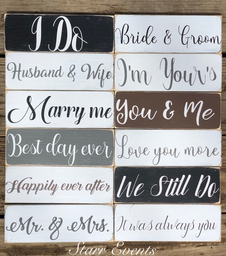 May include: A collection of rectangular wooden signs with white script. Phrases include "I Do", "Bride & Groom", "Husband & Wife", "Marry Me", "Best Day Ever", "Happily Ever After", "Mr. & Mrs.", and more. The signs are in various colors, including black, white, gray, and brown.