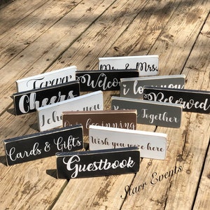 May include: A selection of small, rectangular wooden signs with white lettering on various coloured backgrounds. The signs display phrases such as "Forever," "Welcome," "Cheers," and "Guestbook," suitable for event decoration.