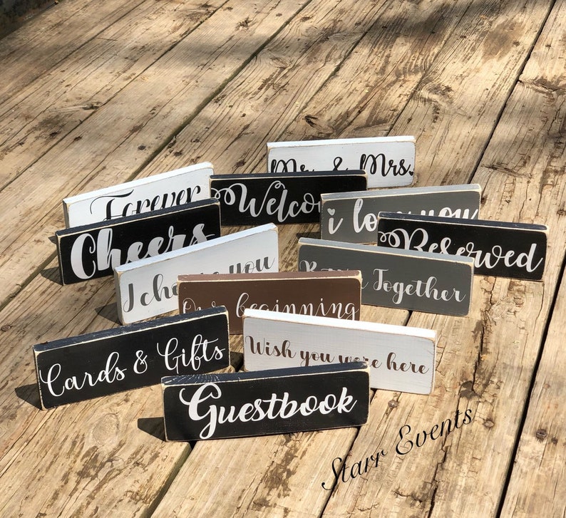 May include: A selection of small, rectangular wooden signs with white lettering on various coloured backgrounds. The signs display phrases such as "Forever," "Welcome," "Cheers," and "Guestbook," suitable for event decoration.