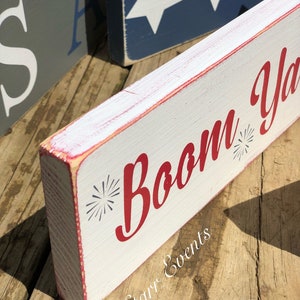May include: A white wooden block sign with the words "Boom Ya" in red script, featuring starburst accents. The sign has red trim and is placed on a wooden surface. Other signs with patriotic themes are visible in the background.