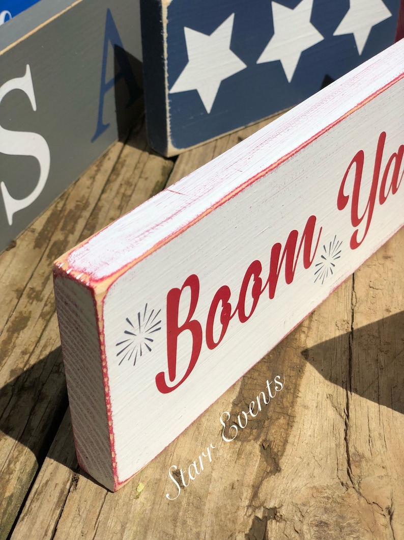 May include: A white wooden block sign with the words "Boom Ya" in red script, featuring starburst accents. The sign has red trim and is placed on a wooden surface. Other signs with patriotic themes are visible in the background.