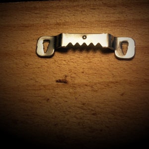 May include: A silver metal picture hanger with a sawtooth edge and keyhole slots for mounting. It's designed for hanging frames or artwork and is shown against a wooden background.