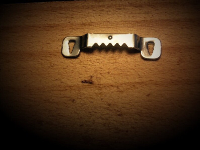 May include: A silver metal picture hanger with a sawtooth edge and keyhole slots for mounting. It's designed for hanging frames or artwork and is shown against a wooden background.