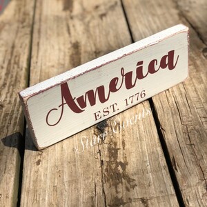 May include: A rectangular wooden sign with the word "America" in a burgundy script font. Below the word is "EST. 1776" in a smaller, block font. The sign has a distressed, vintage look with a cream-coloured background and red trim.