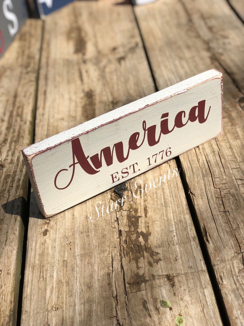 May include: A rectangular wooden sign with the word "America" in a burgundy script font. Below the word is "EST. 1776" in a smaller, block font. The sign has a distressed, vintage look with a cream-coloured background and red trim.