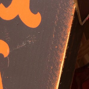 May include: Close-up of a section of a sign with orange lettering on a dark brown background. The edge of the sign has an orange border with a decorative pattern. The sign appears to be made of wood.