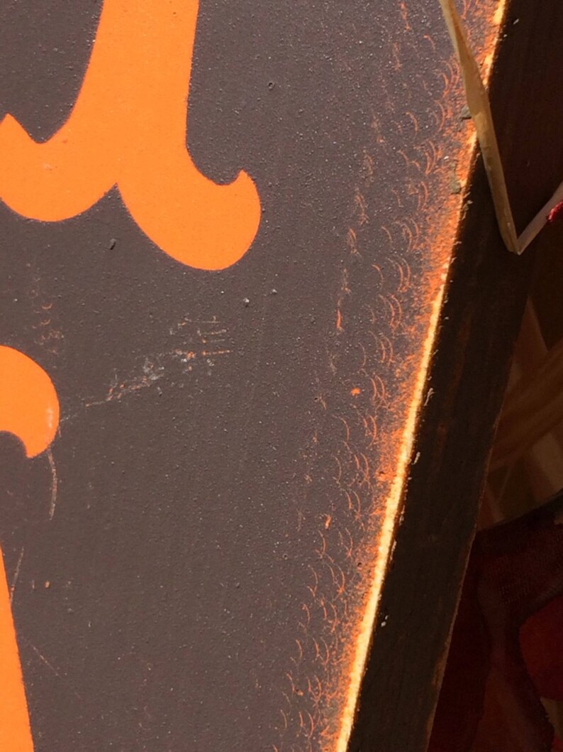May include: Close-up of a section of a sign with orange lettering on a dark brown background. The edge of the sign has an orange border with a decorative pattern. The sign appears to be made of wood.