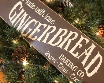 Puede incluir: Un letrero de madera marrón con letras blancas que dice "Made with love GINGERBREAD BAKING CO. Bread Cake Cookies." El letrero está rodeado de ramas de pino verdes, piñas y bayas rojas.